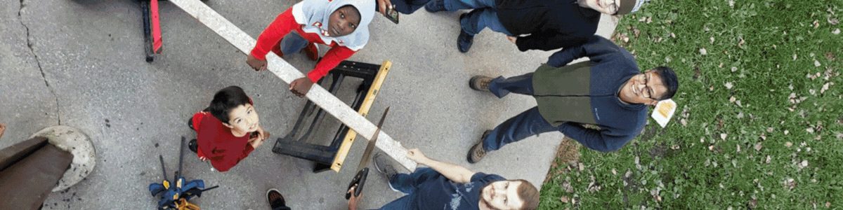 Seen from above, students and an instructor carry a long board across a yard during a build.