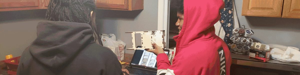 A student holds up a freshly cut plywood piece next to a laptop showing the design it was cut from.